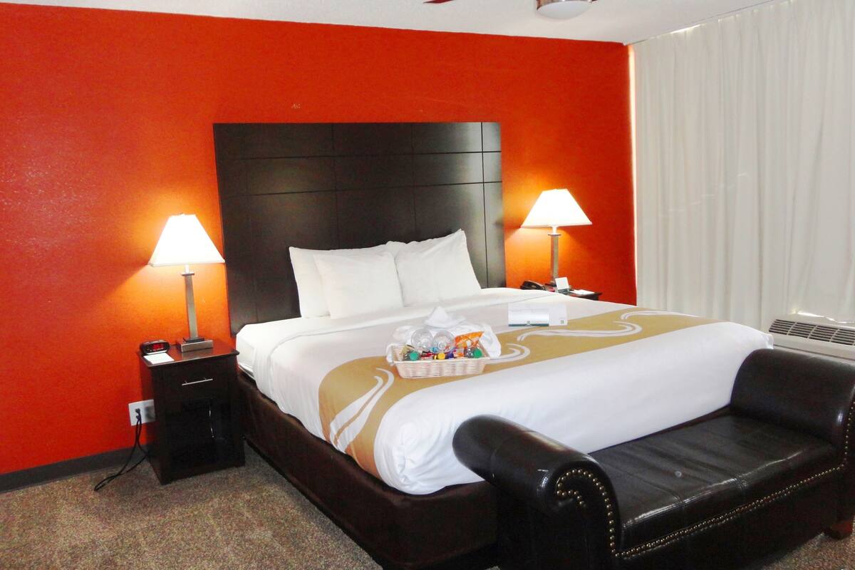 A hotel room with a large bed, two bedside tables with lamps, a ceiling fan, a black leather armchair, and a window with white curtains.