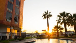 A swimming pool with a sunset in the background, surrounded by palm trees and a building with multiple windows.