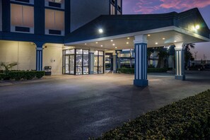 Entrada de un hotel con un vestíbulo cubierto, puertas de vidrio y una palmera al frente.