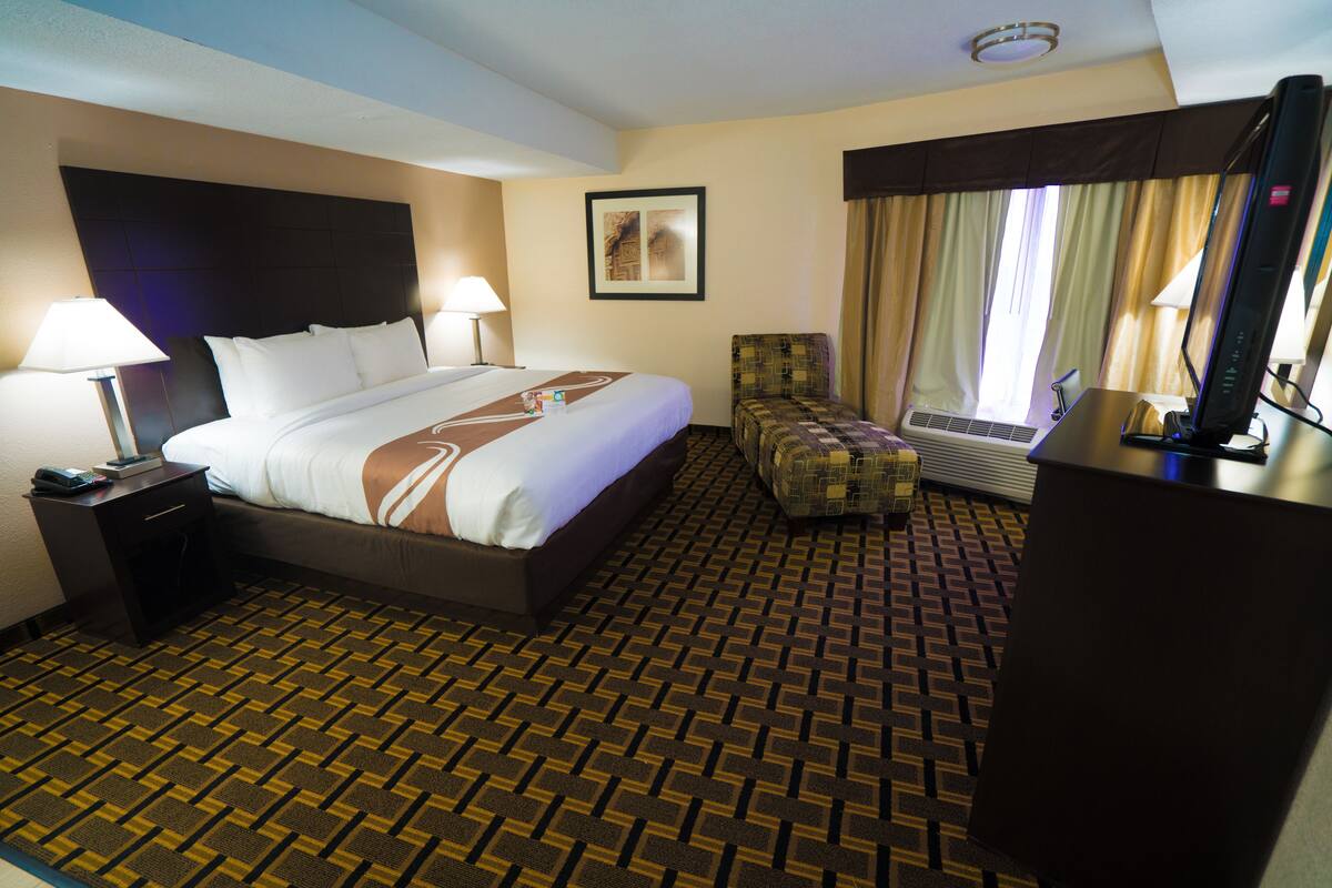 A hotel room with a large bed, two bedside tables with lamps, a chair, a desk with a computer, and a window with curtains.