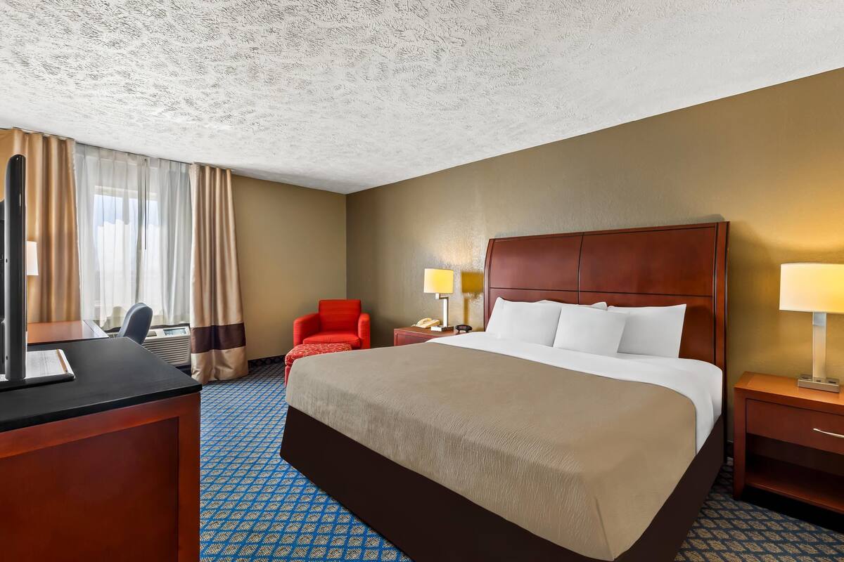 Quarto de hotel com uma cama grande, uma escrivaninha, uma cadeira e uma mesa de cabeceira com luminárias.
