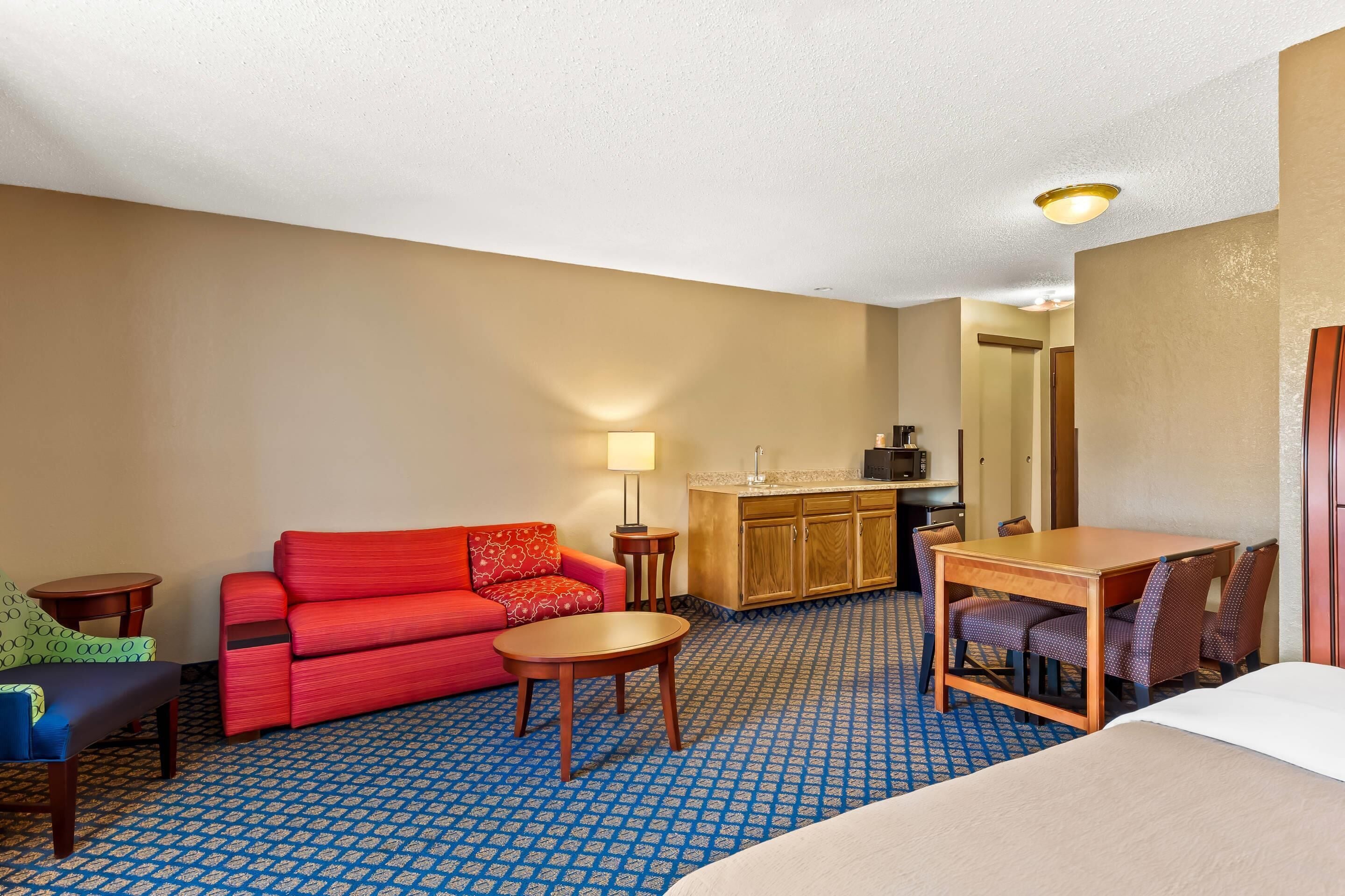 A hotel room with a red sofa, a small round table, a wooden desk with chairs, a bed, and a kitchenette area.