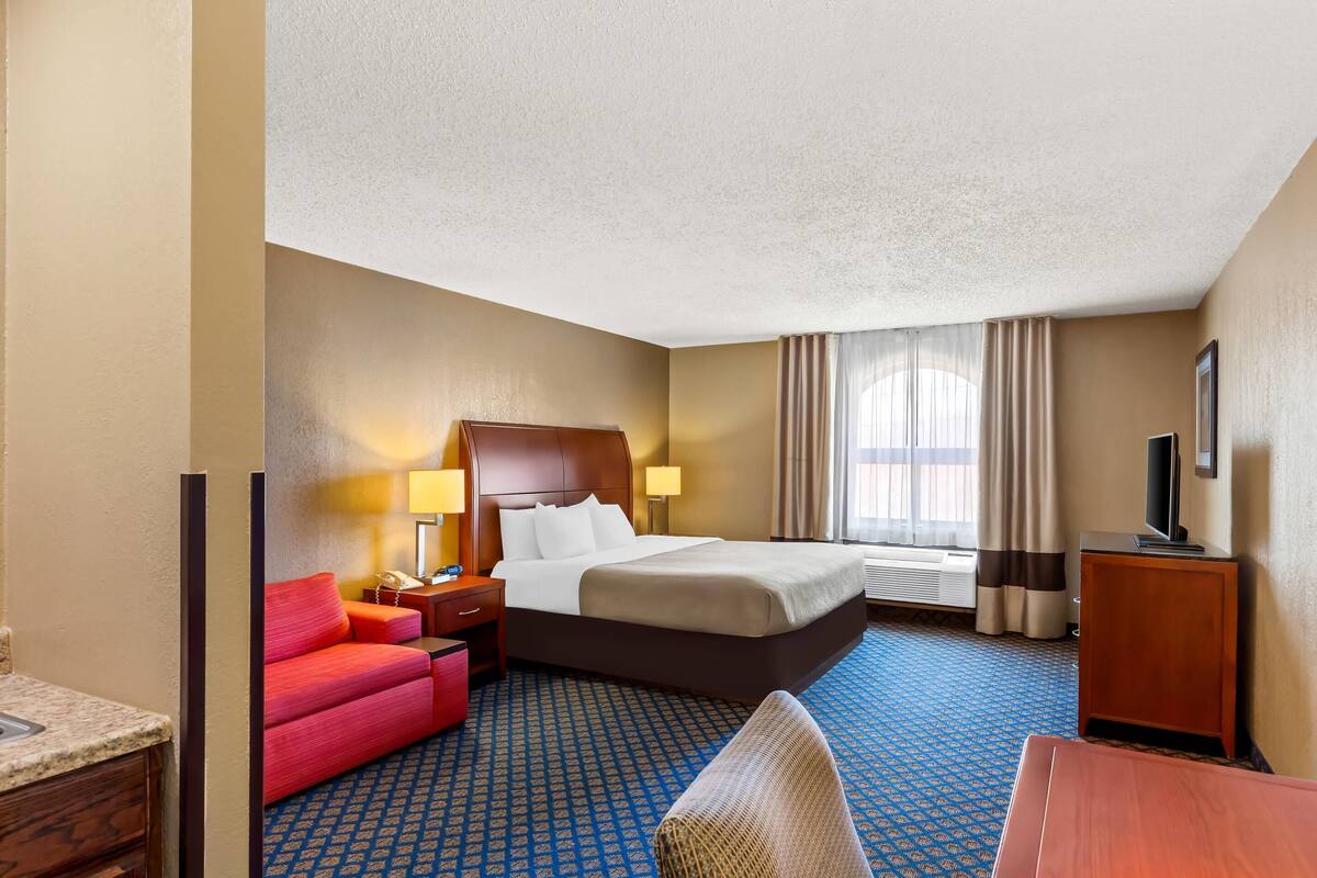 Quarto de hotel com cama, escrivaninha, cadeira, sofá, televisão e janela com cortinas.