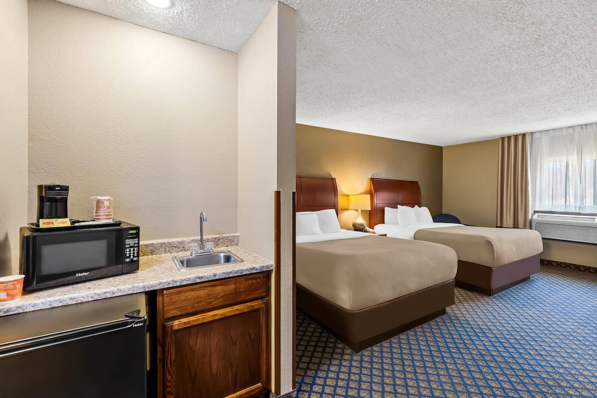 Quarto de hotel com cozinha compacta, duas camas, banheiro e janela com cortinas.