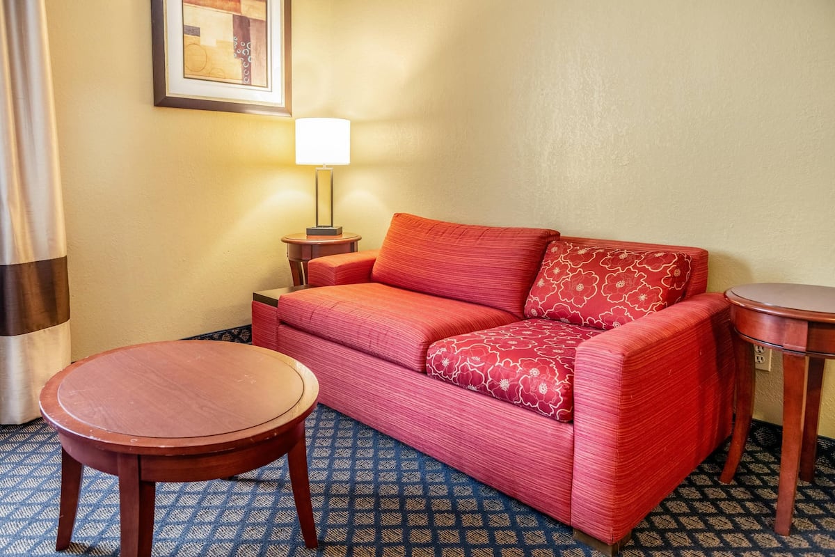 Quarto de hotel com um sofá vermelho, uma pequena mesa de madeira, uma lâmpada e um tapete estampado.