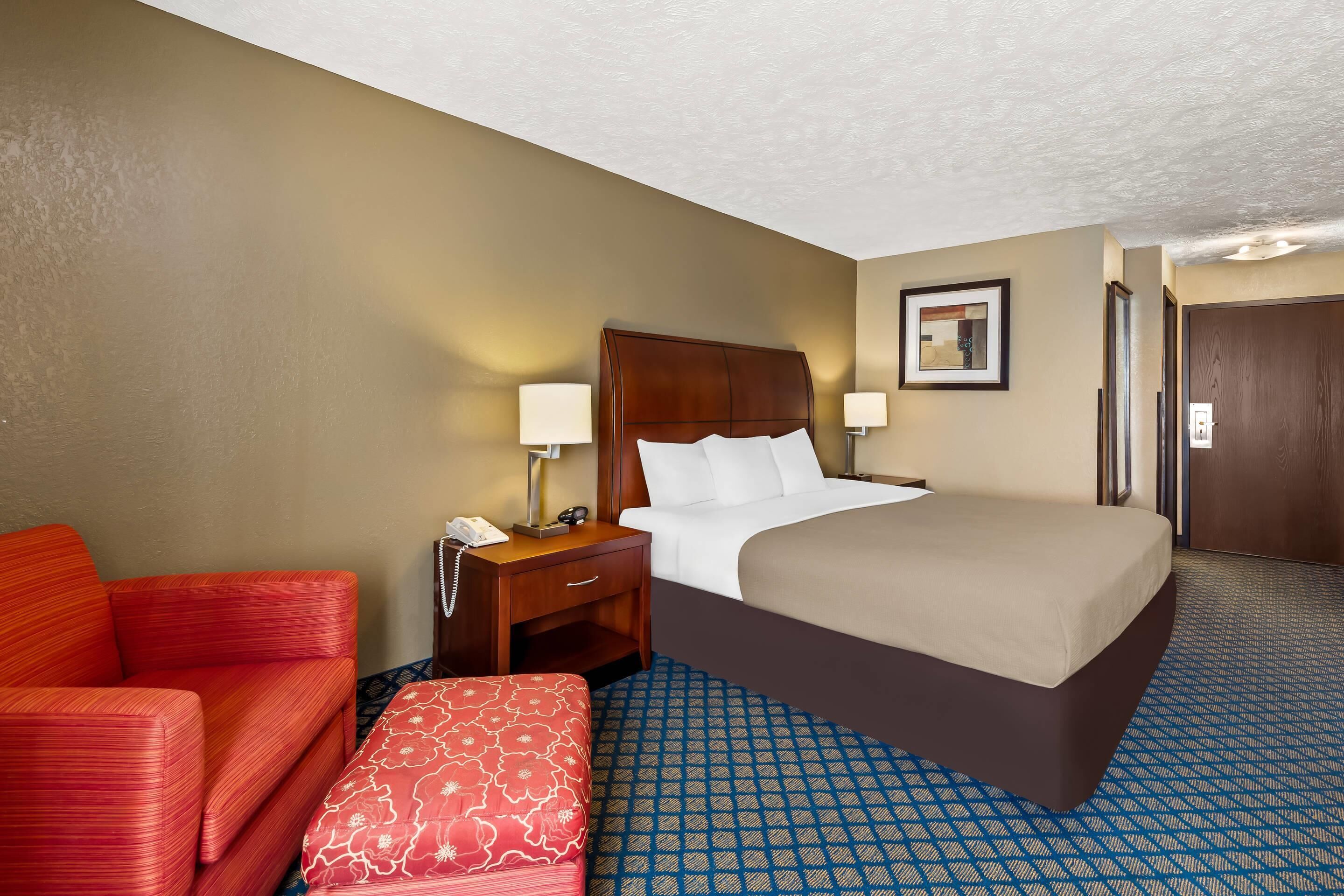A hotel room with a bed, a nightstand, a red sofa, a patterned ottoman, and a patterned carpet.