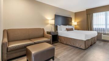 A hotel room with a bed, a sofa, an ottoman, a nightstand, and a lamp.