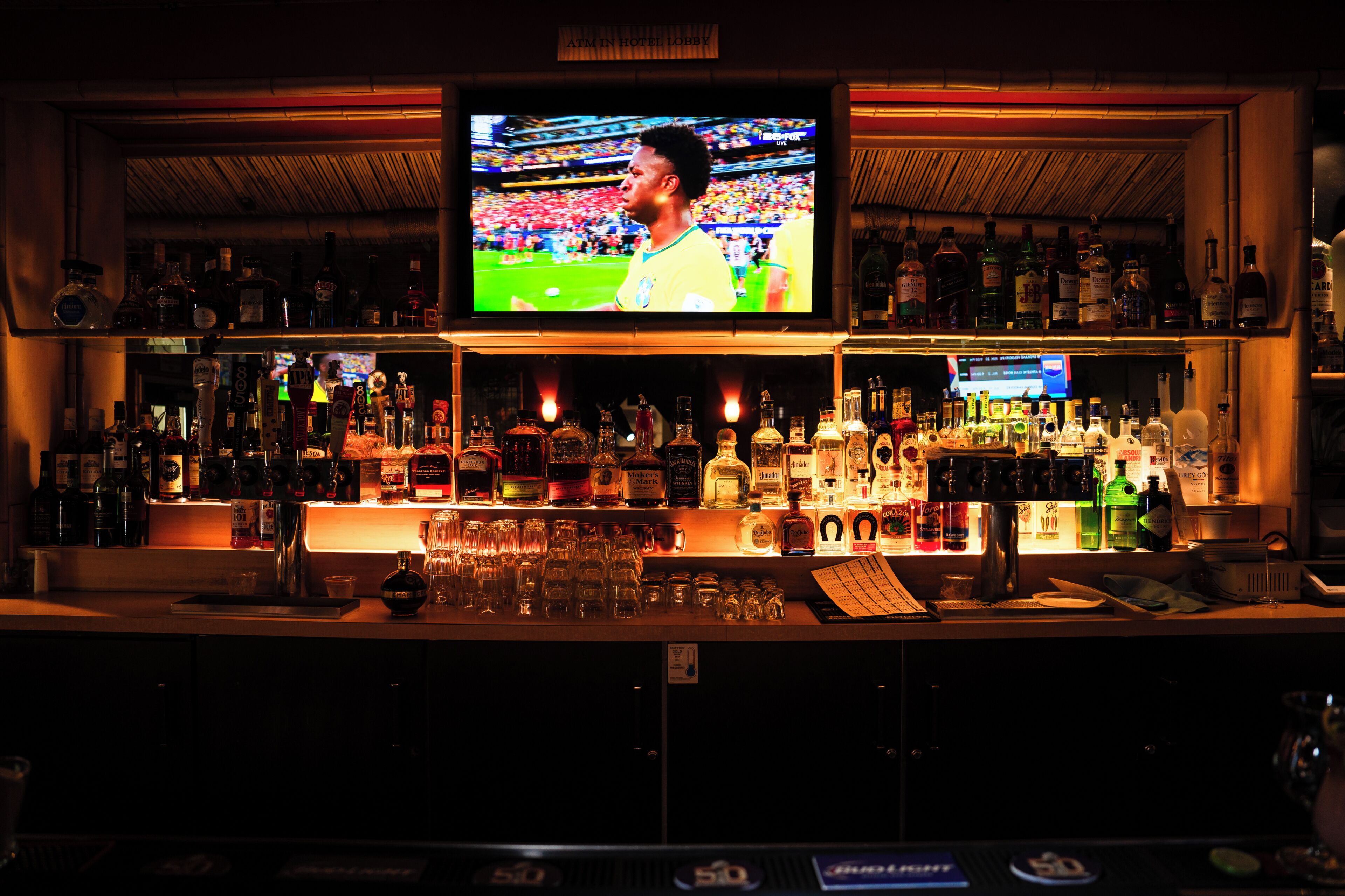 A bar with a television displaying a sports event, shelves stocked with various liquor bottles, and a bar counter with glasses and a cash register.