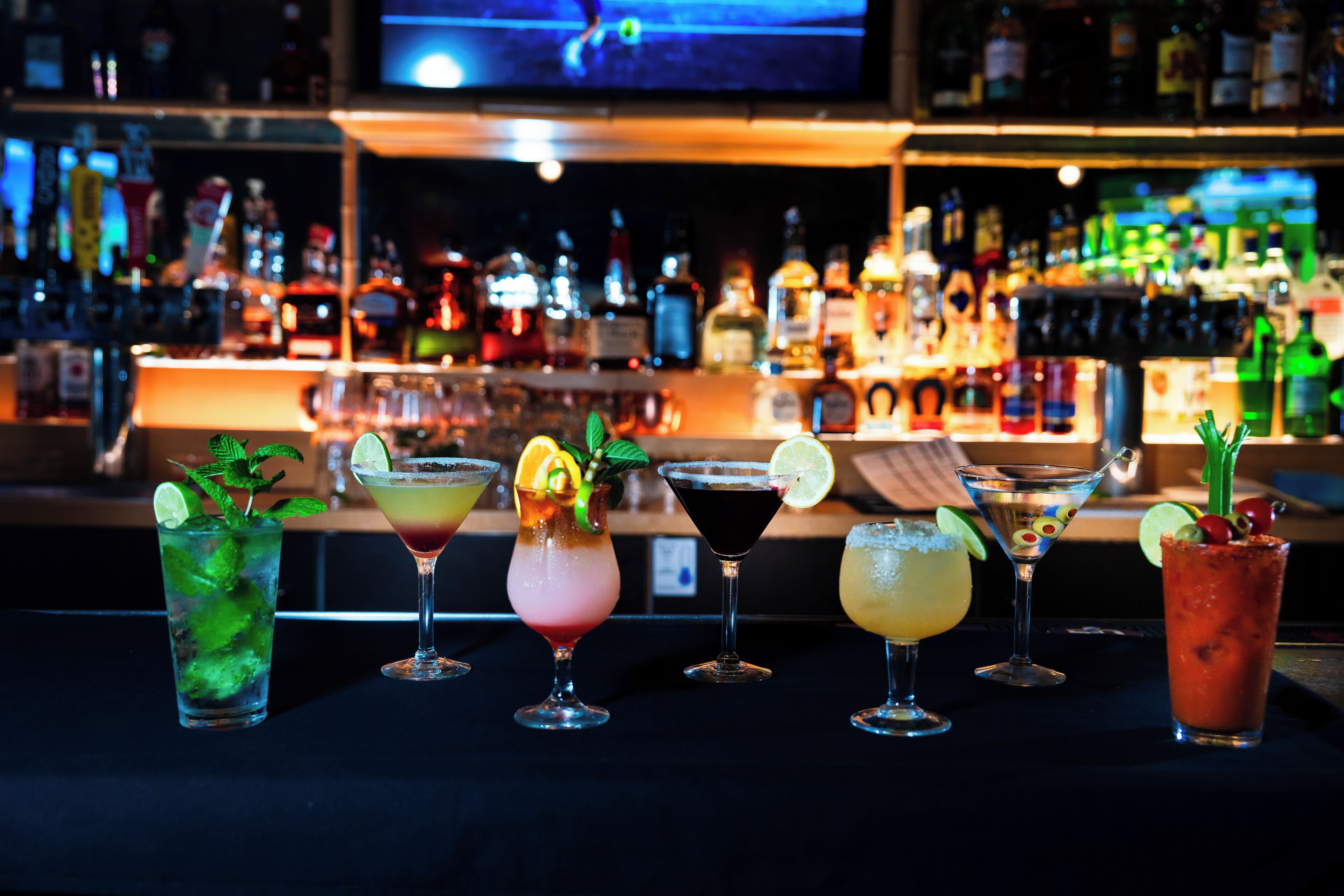 A bar with various cocktails, a television screen in the background, and shelves of bottles.