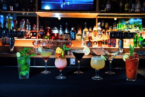 Eine Bar mit verschiedenen Cocktails, einem Fernseher im Hintergrund und Regalen voller Flaschen.