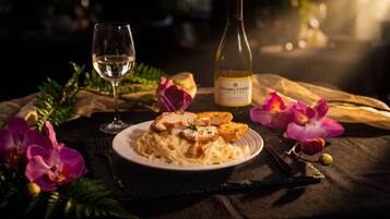 Un plato de pasta con pollo a la parrilla y pan tostado, una copa de vino blanco y una botella de vino Sonoma Creek sobre una mesa con orquídeas y un paño oscuro.