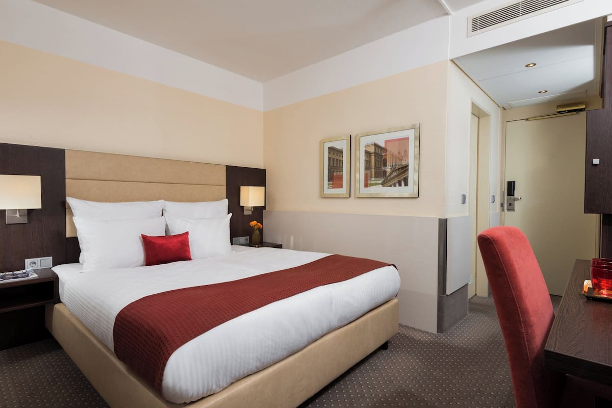 A hotel room with a large bed, two bedside tables with lamps, a desk with a chair, and framed pictures on the wall.