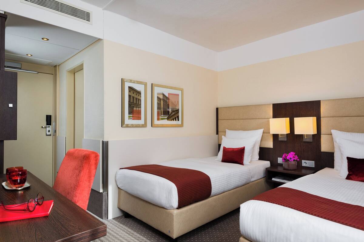 A hotel room with two beds, a desk, and a chair. There are framed pictures on the wall, a nightstand with a lamp, and a vase with flowers.