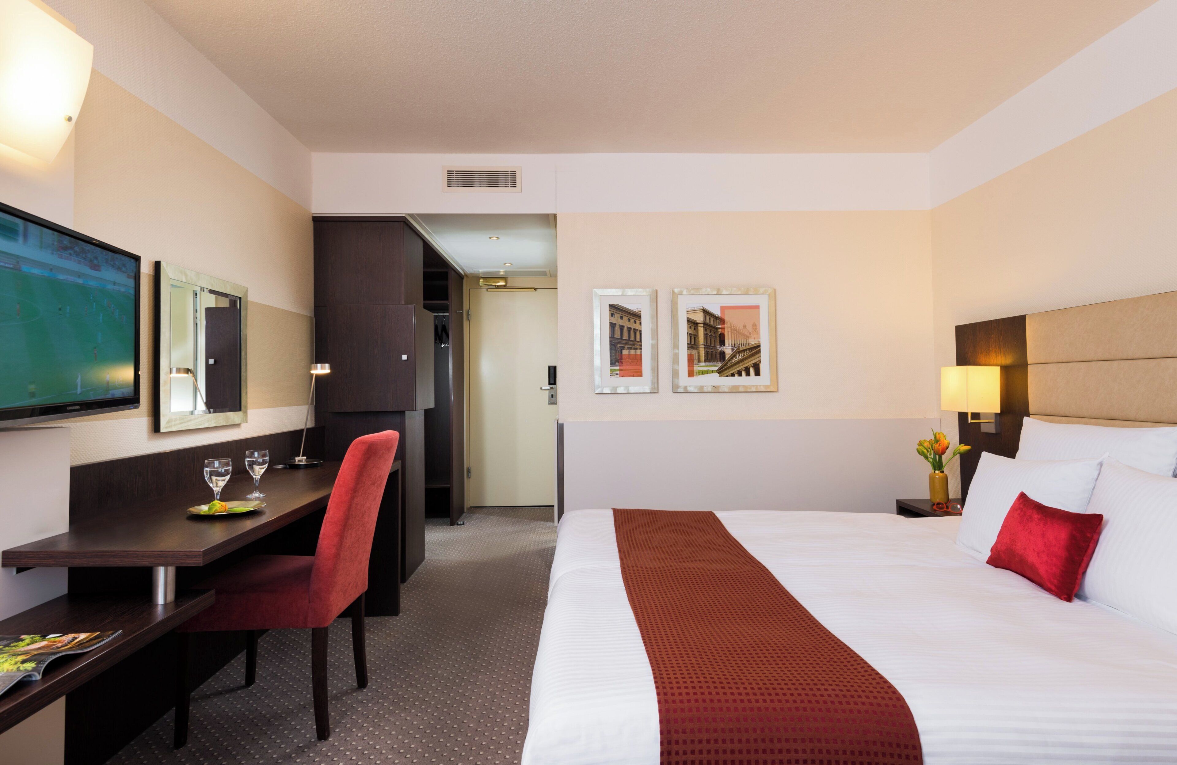 Habitación de hotel con cama, escritorio con silla, televisor y cuadros en la pared.