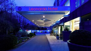 El exterior del Hotel Leonardo, con un acceso cubierto, una zona de recepción y un camino que lleva al edificio.