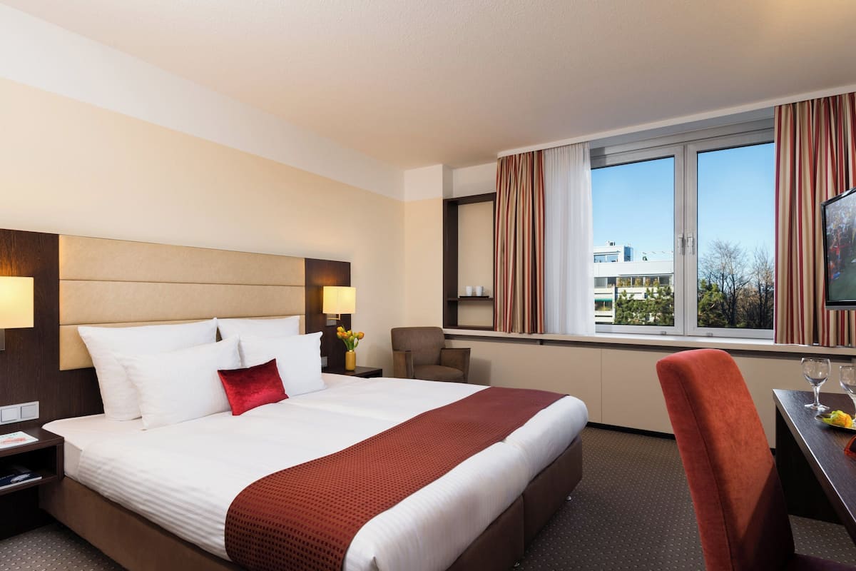 A hotel room with a large bed, a desk with a chair, a TV, and a window with curtains.