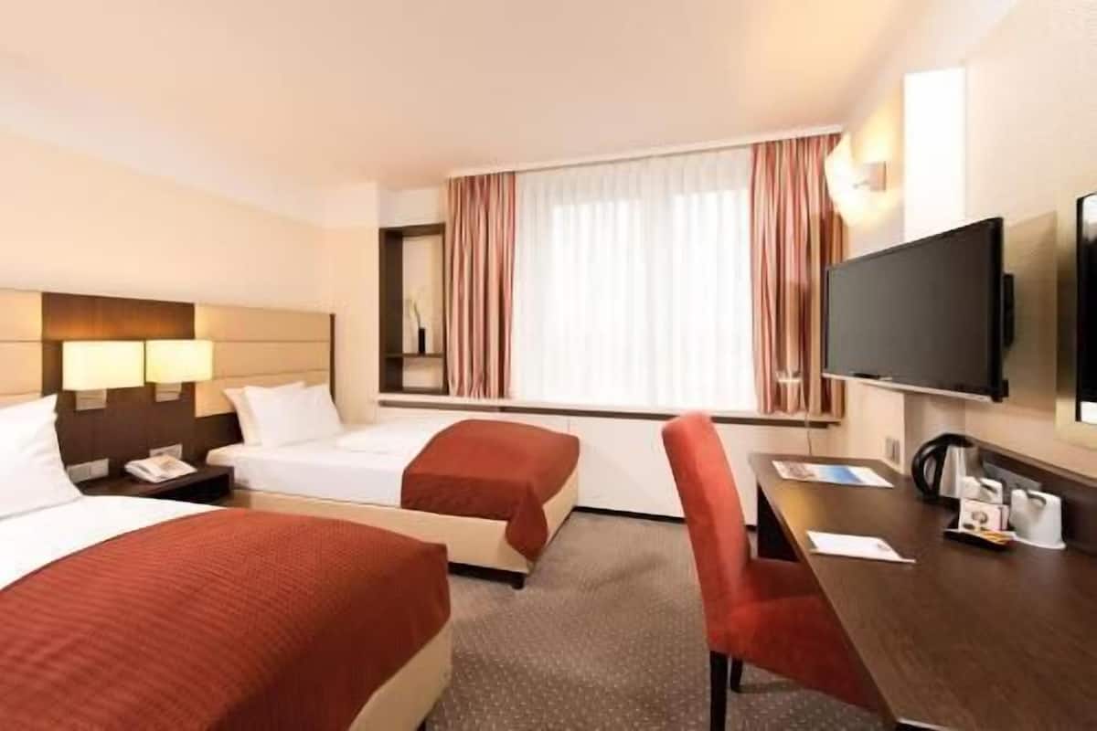 A hotel room with two beds, a desk, a television, and a window with curtains.