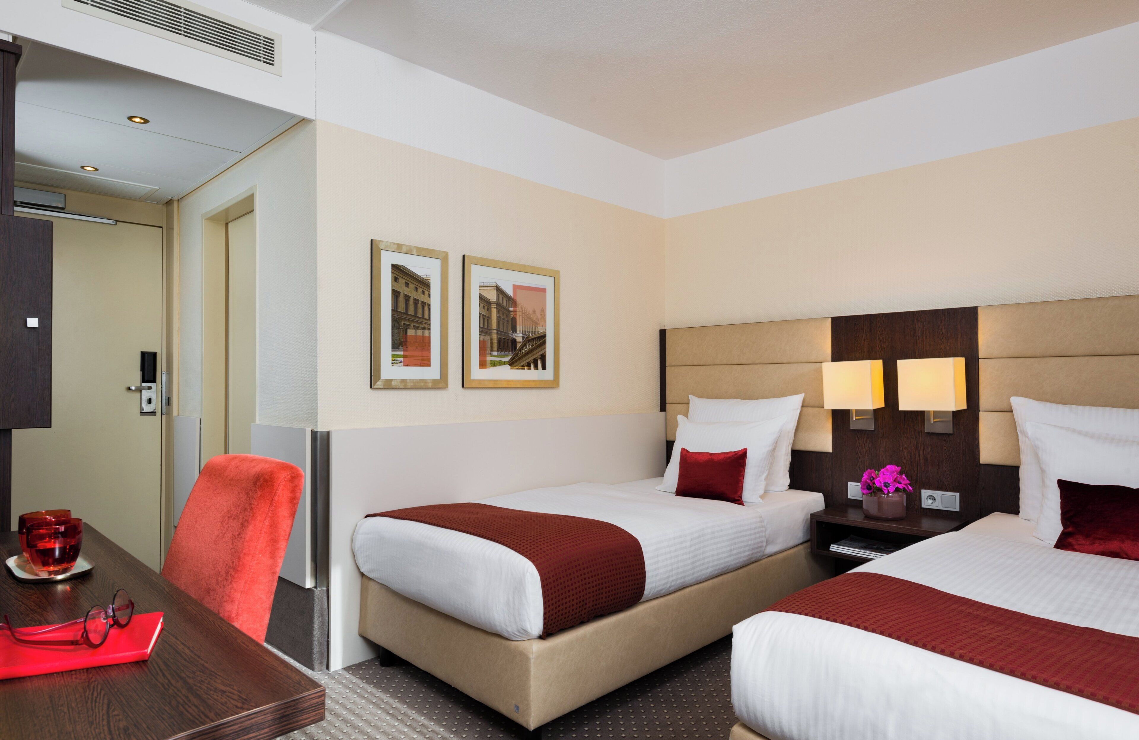 A hotel room with two beds, a desk, and a chair. There are framed pictures on the wall, a nightstand with a lamp, and a vase with flowers.