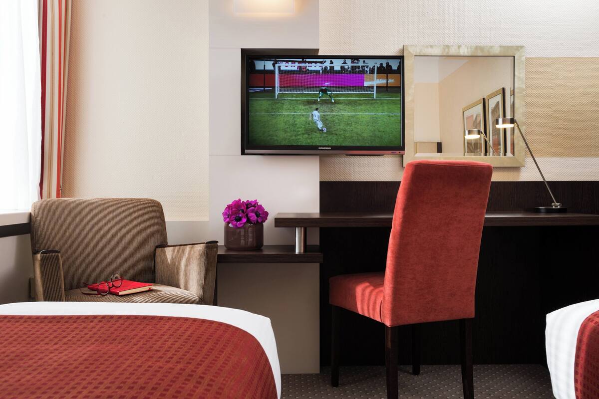 A hotel room with a bed, a desk with a TV, a chair, and a small table with a vase of flowers.