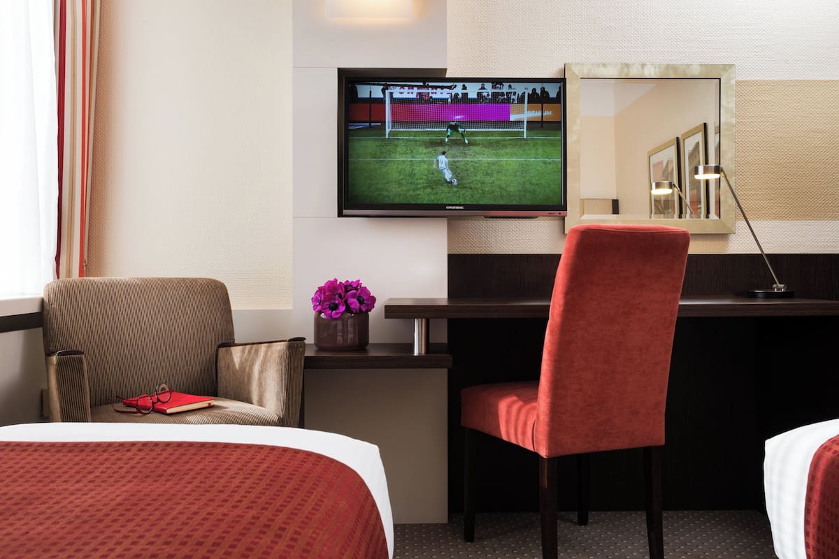 Une chambre d’hôtel avec un lit, un bureau avec une télévision, une chaise et une petite table sur laquelle est posé un vase de fleurs.