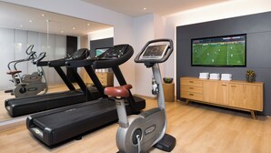 Un gimnasio moderno en casa con cinta de correr, elíptica y bicicleta estática, un espejo grande y un televisor que muestra un partido de fútbol.