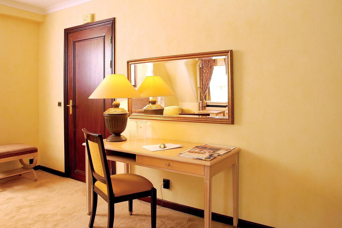 A hotel room with a desk, two lamps, a chair, a bench, and a door.