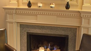 A hotel room with a fireplace, two chairs, a round table, and a painting on the wall.