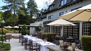 An outdoor dining area with multiple tables and chairs, surrounded by large umbrellas, and a building with large windows in the background.