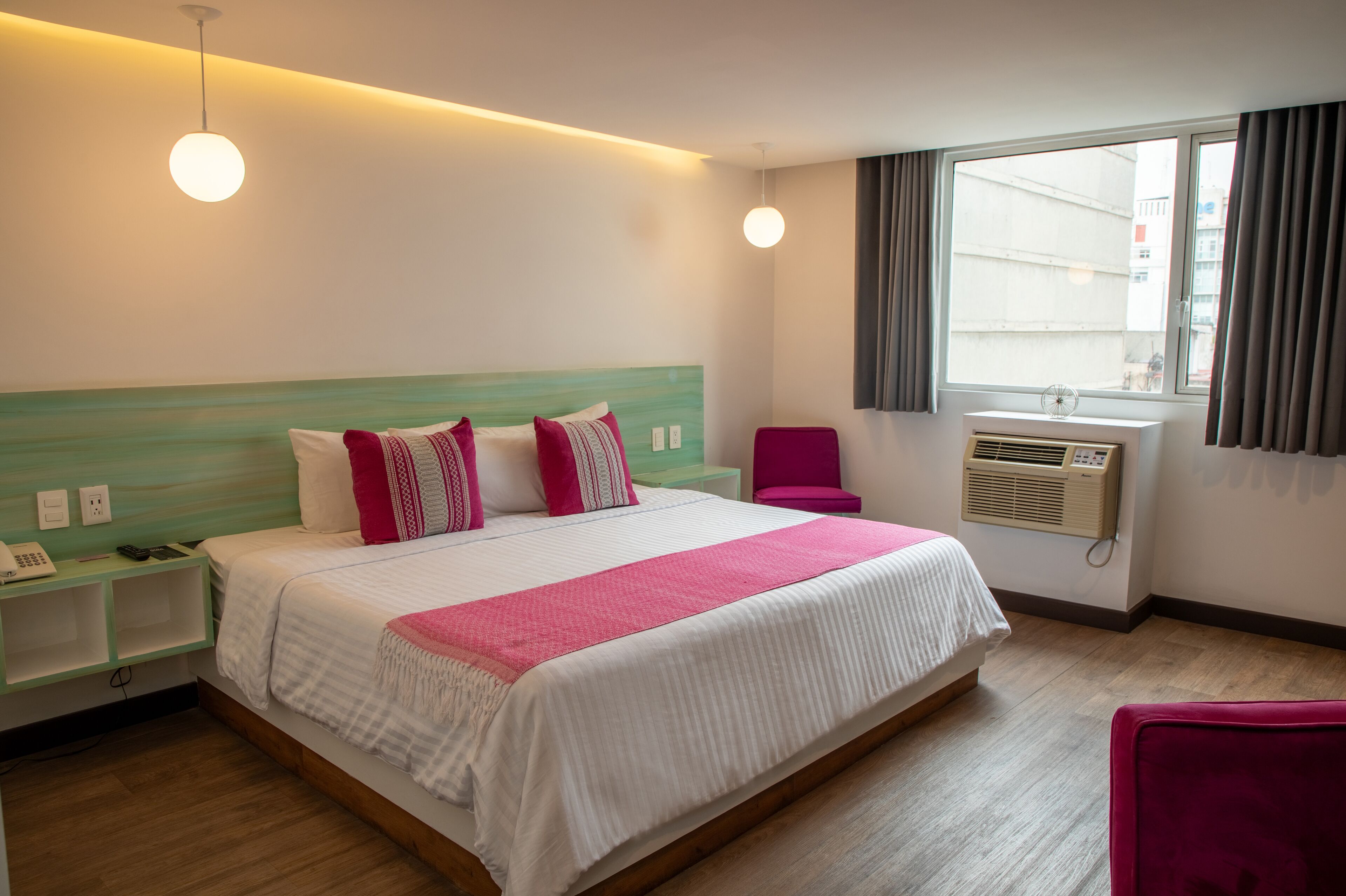 Una habitación de hotel con una cama grande, una mesita de noche, una silla, una ventana con cortinas y un equipo de aire acondicionado.