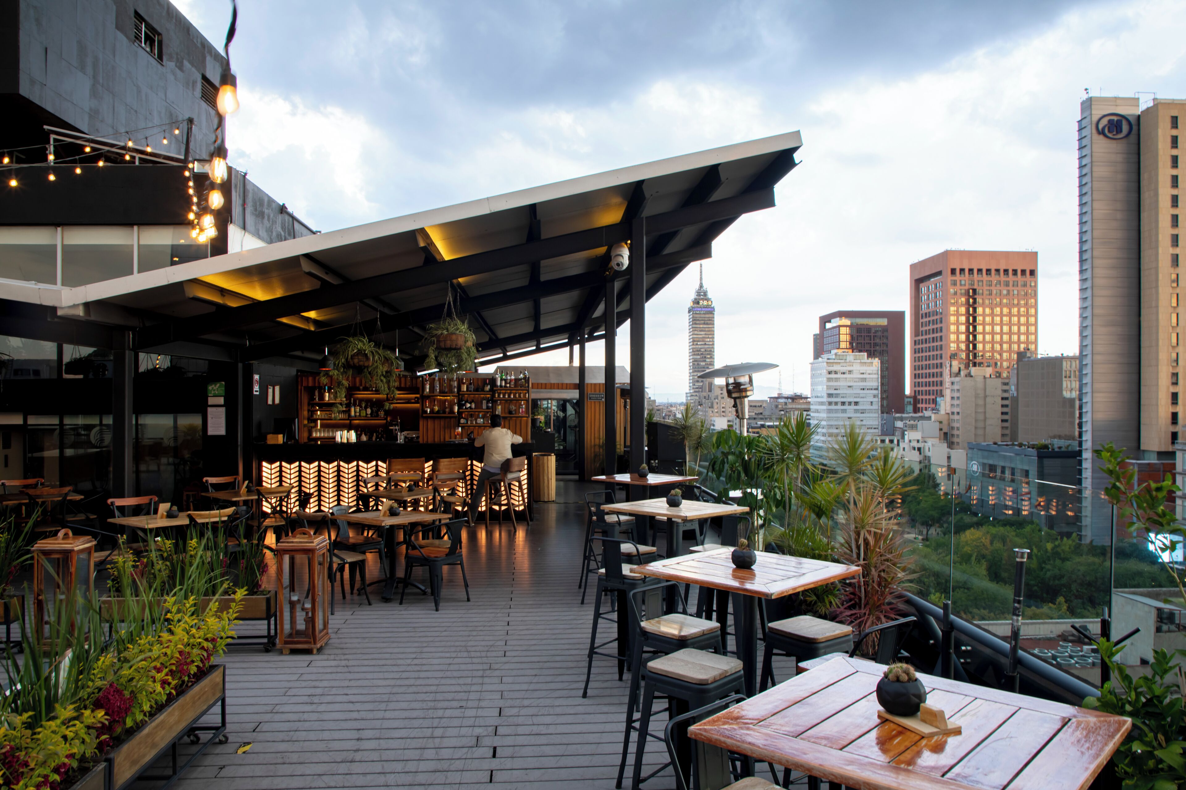 An outdoor seating area with tables and chairs, a bar with shelves of bottles, and a city skyline in the background.