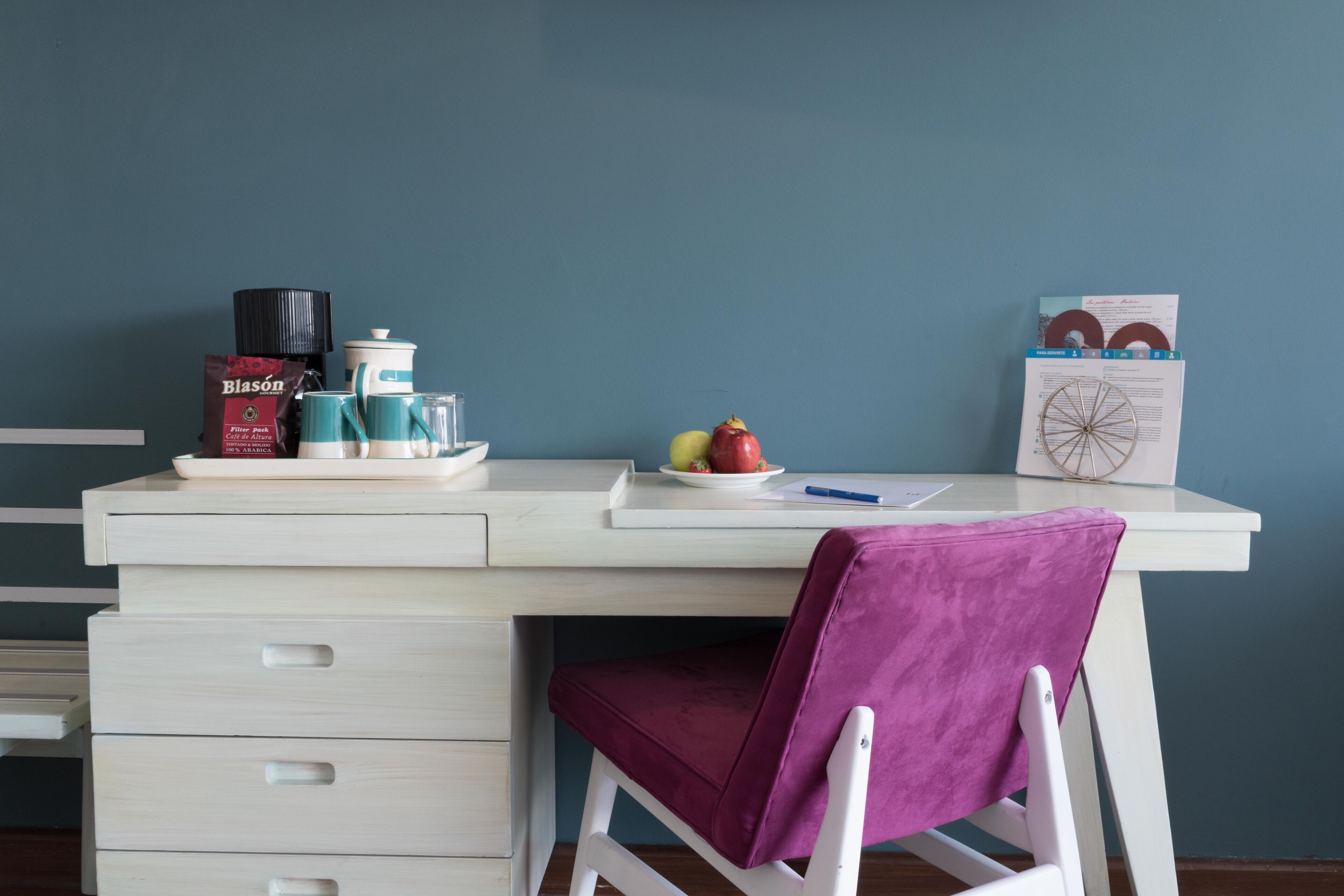 Un escritorio con una silla, una cafetera, un juego de té, un plato con fruta y un calendario.