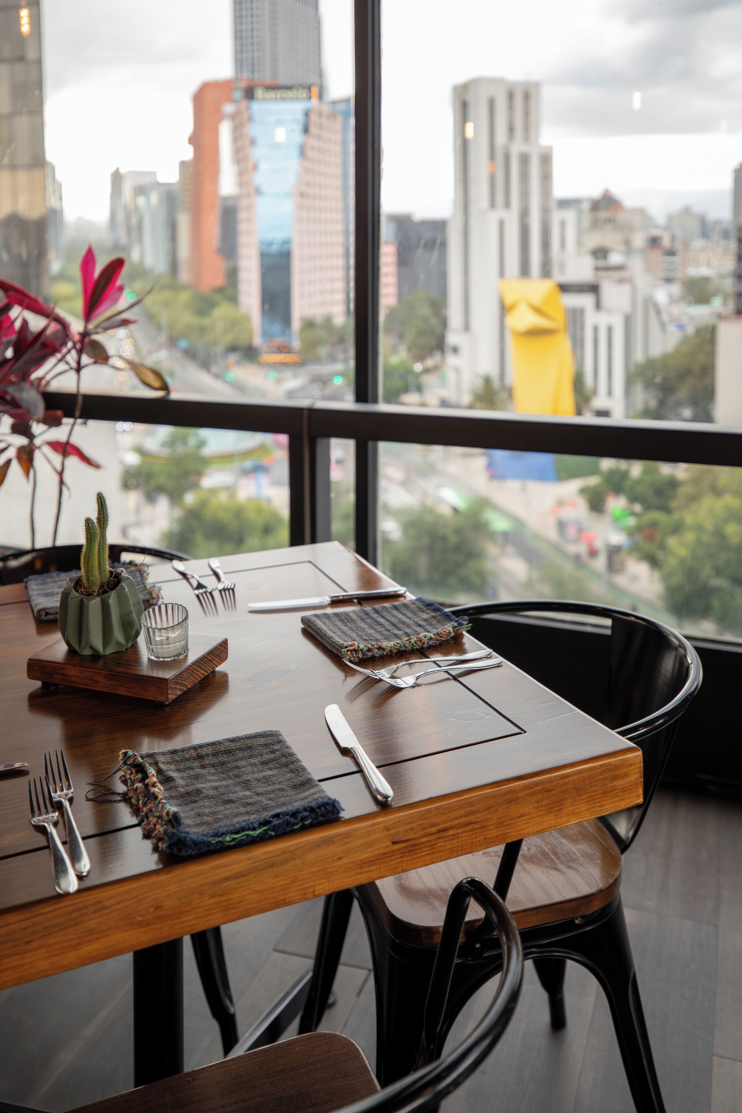 Mesa lista con cubiertos y una planta de cactus, con vista a un paisaje urbano a través de un amplio ventanal.