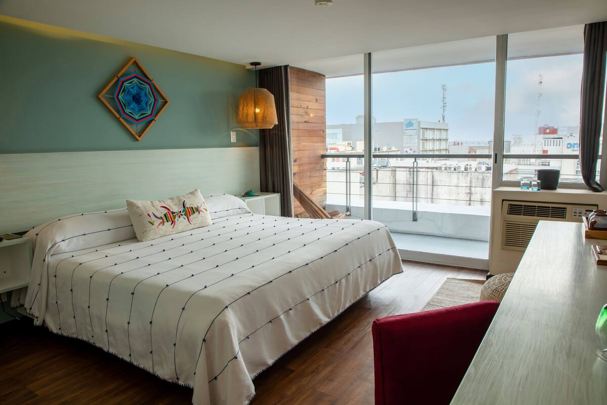A hotel room with a large bed, a wooden headboard, a view of the city through large windows, and a balcony with a railing.