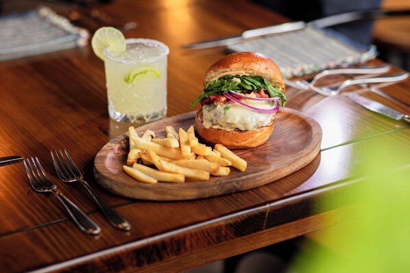 Hamburguesa con papas fritas sobre una tabla de madera, una bebida con una cuña de lima y cubiertos sobre la mesa.