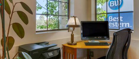 Un espacio de trabajo con computadora, impresora y lámpara cerca de una ventana que da a árboles.
