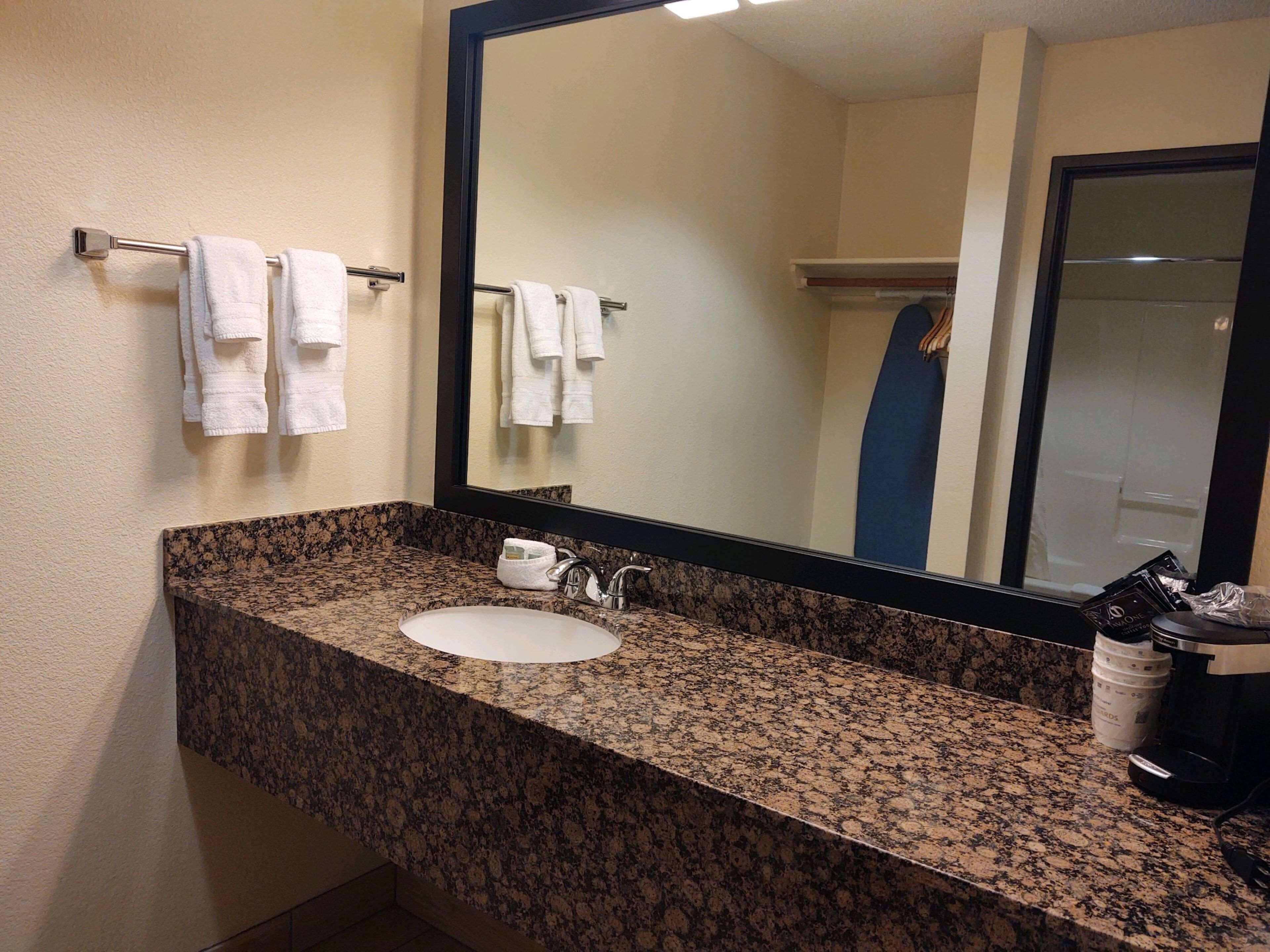 Une salle de bain avec un comptoir en granit, un lavabo blanc et un grand miroir. Des serviettes sont suspendues sur un porte-serviettes placé au-dessus du lavabo.