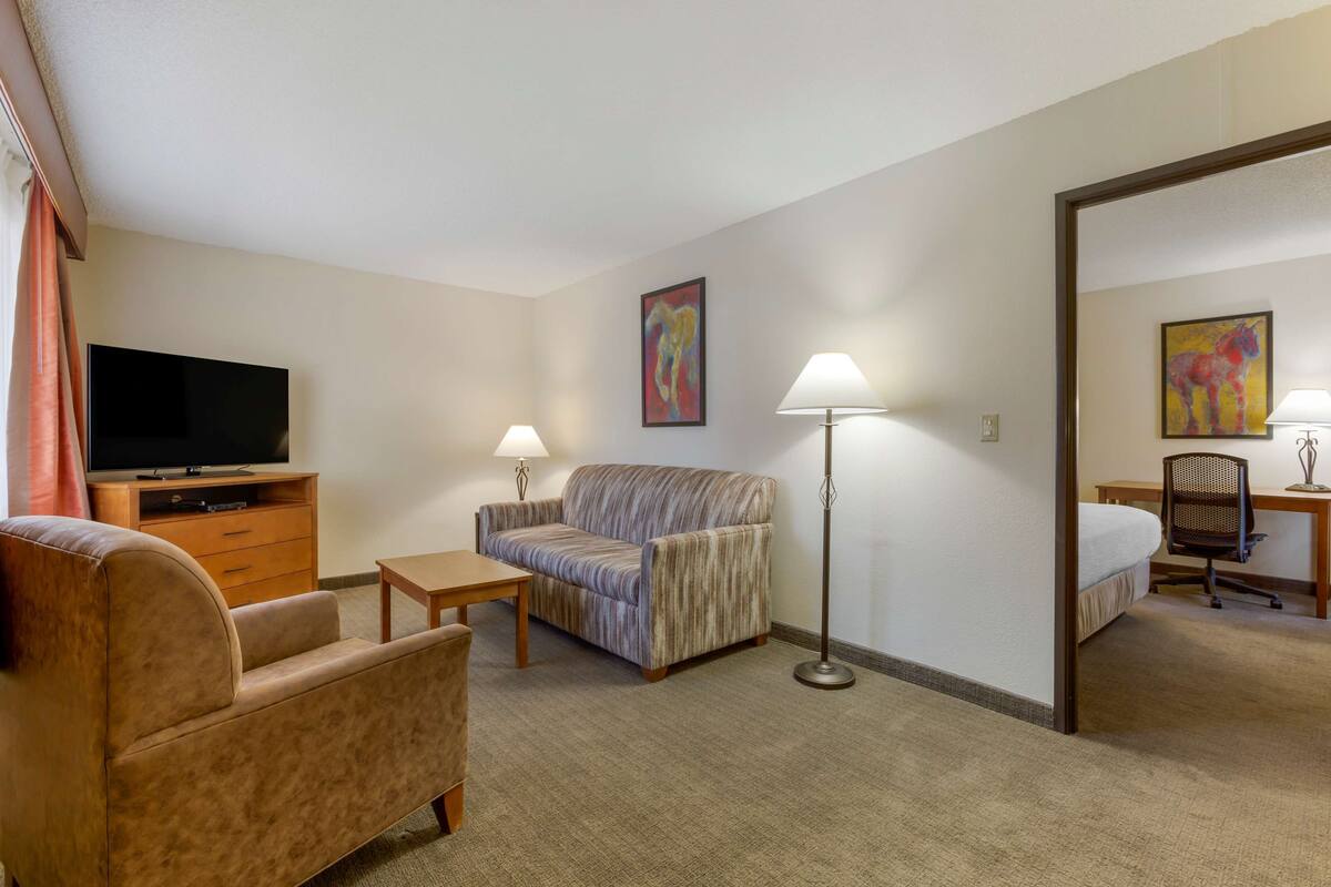 A hotel room with a sofa, armchair, TV, desk, lamp, and artwork.