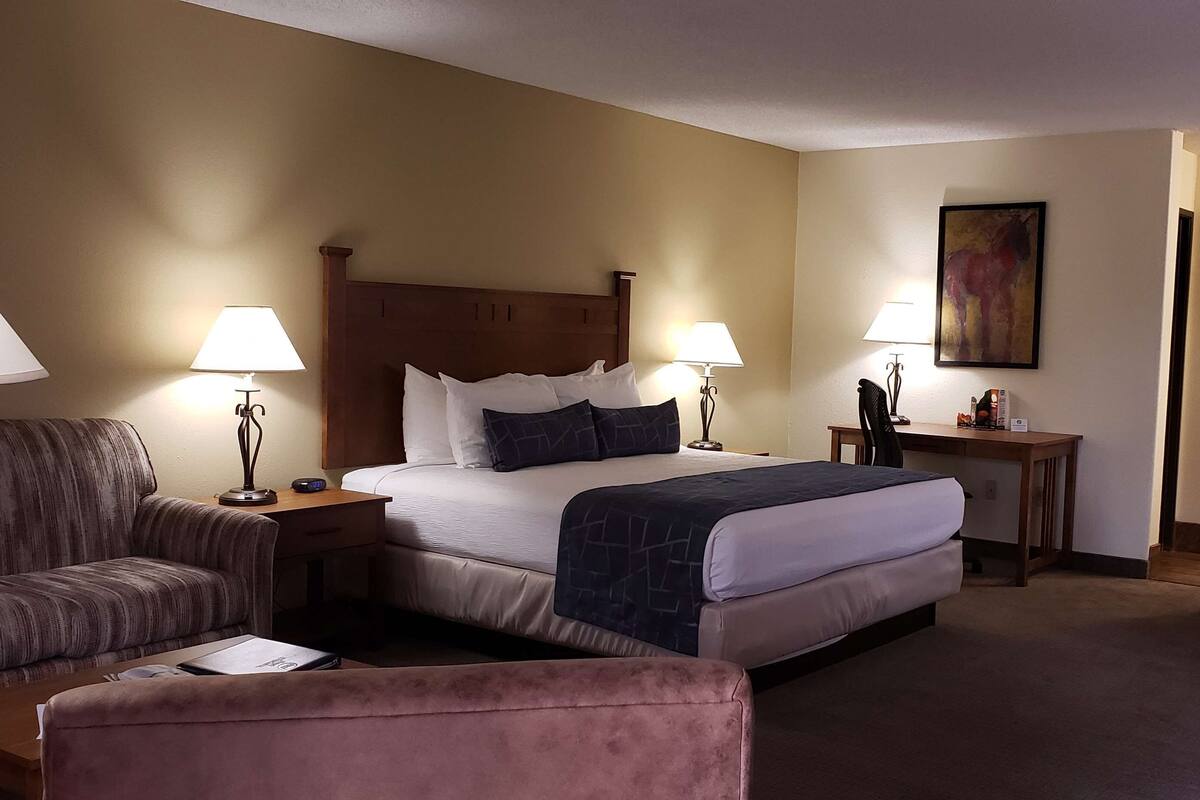A hotel room with a bed, two bedside tables with lamps, a desk, a chair, a couch, and a wooden headboard with a built-in TV.