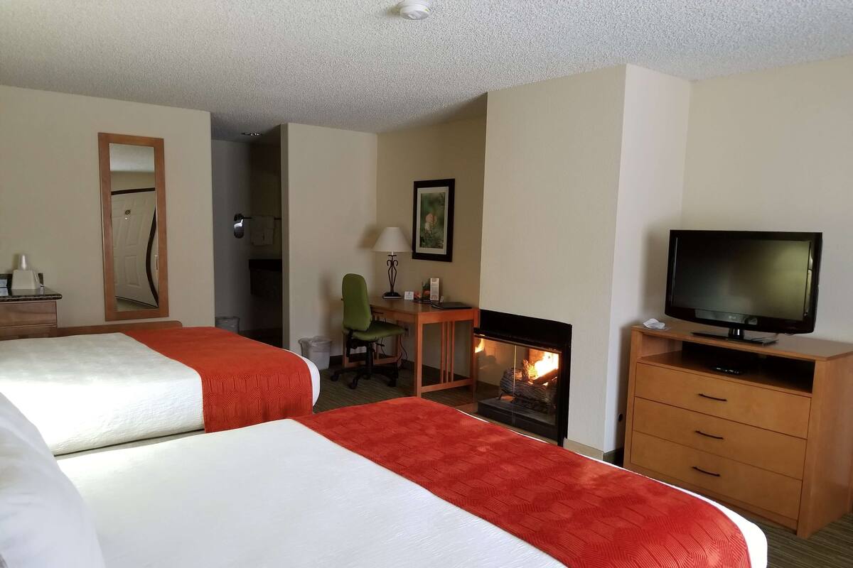 A hotel room with two beds, a television, a desk with a chair, and a fireplace.