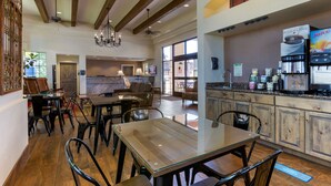 Un comedor amplio con una mesa y sillas de vidrio, una zona de cocina con máquinas de café, y un gran ventanal que proporciona luz natural.