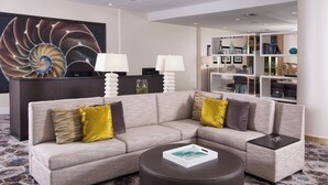 A modern hotel lobby with a grey sectional sofa, a round ottoman, and a reception desk with two lamps. - Courtyard Miami in Coral Gables by Marriott (Coral Gables)