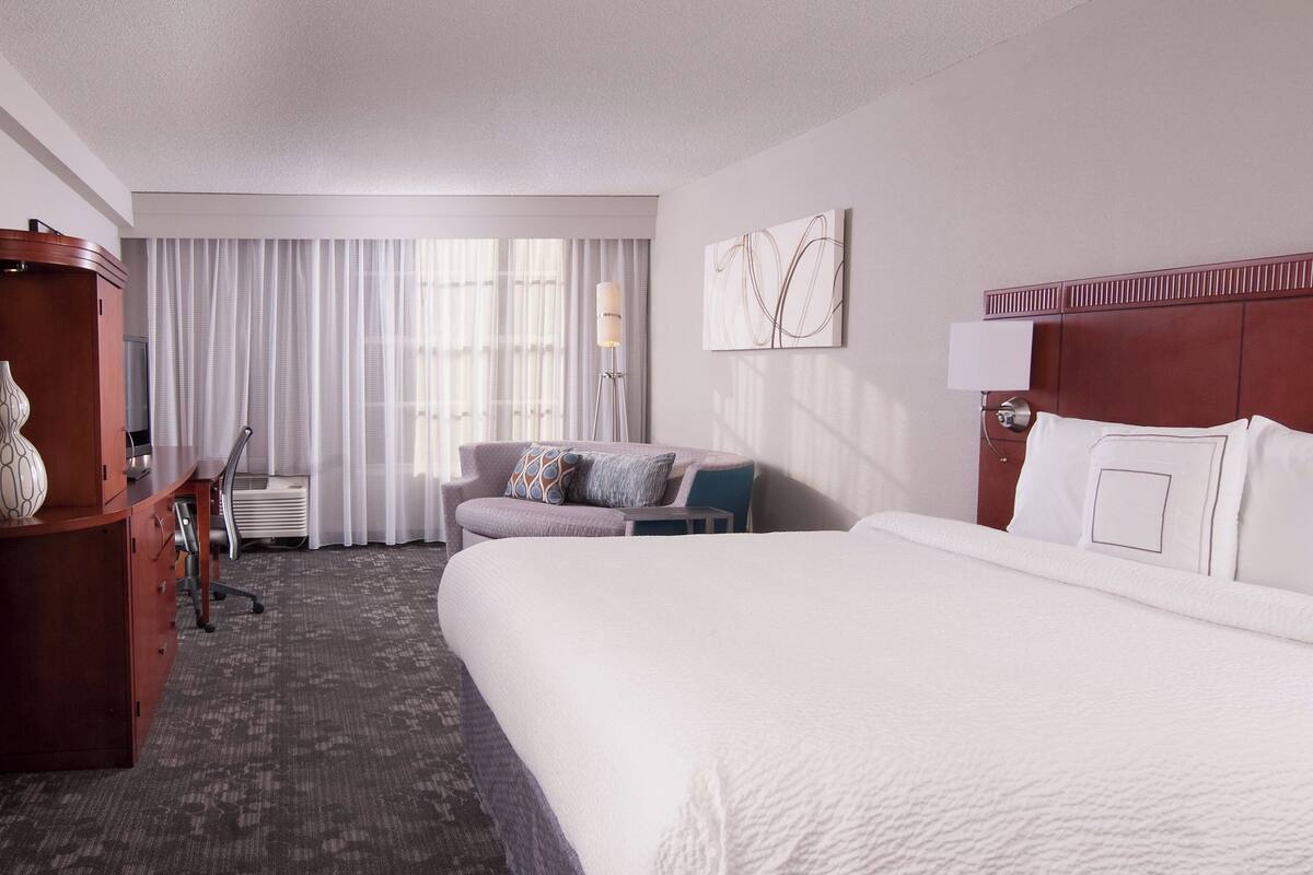 Una habitación de hotel con una cama grande, un escritorio con silla, un sofá y un ventanal con cortinas.