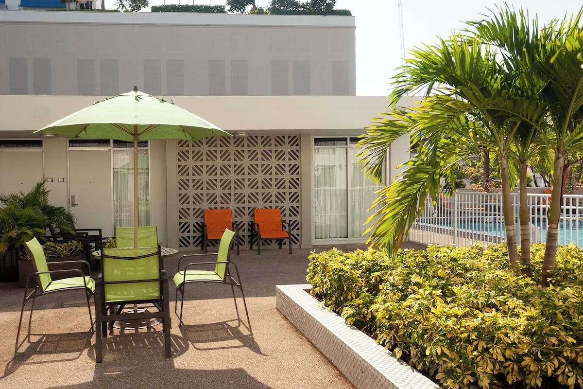 Un área de estar al aire libre con una mesa y sillas, una sombrilla verde y una piscina al fondo.