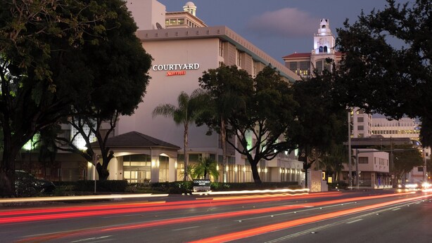 A Courtyard Marriott hotel with a prominent clock tower, surrounded by trees and modern buildings. - Courtyard Miami in Coral Gables by Marriott (Coral Gables)