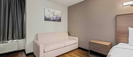 A hotel room with a sofa, a bedside table, a headboard, and a wall-mounted artwork.