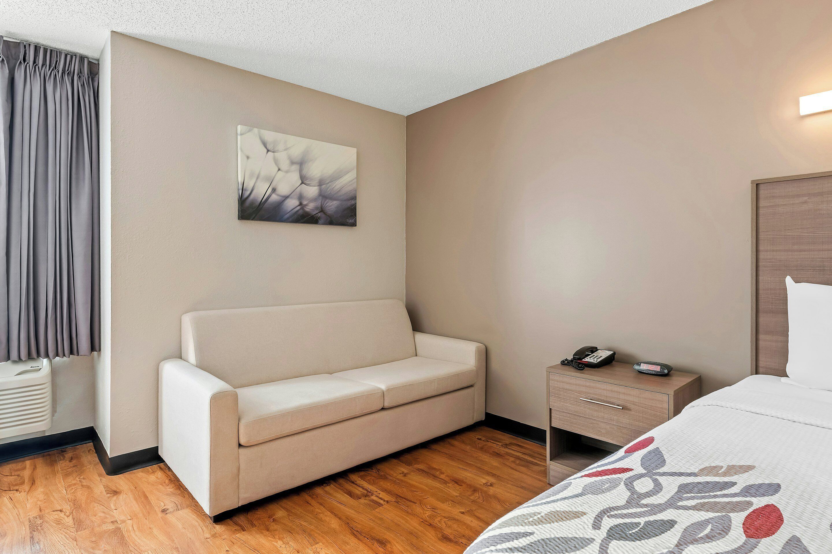 A hotel room with a beige sofa, a bedside table with a phone, a wooden floor, and a painting on the wall.
