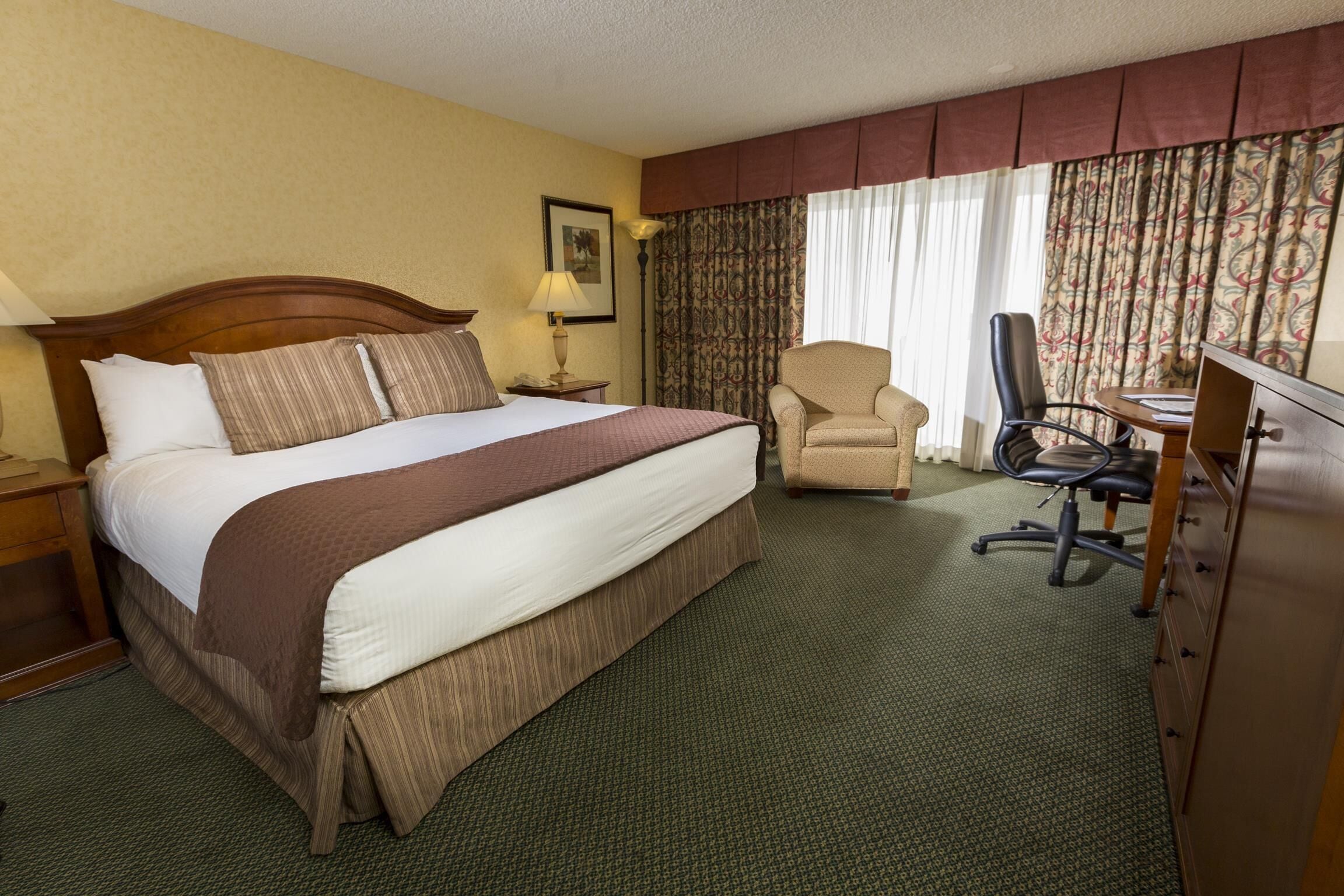 A hotel room with a large bed, a desk, a chair, and a dresser.