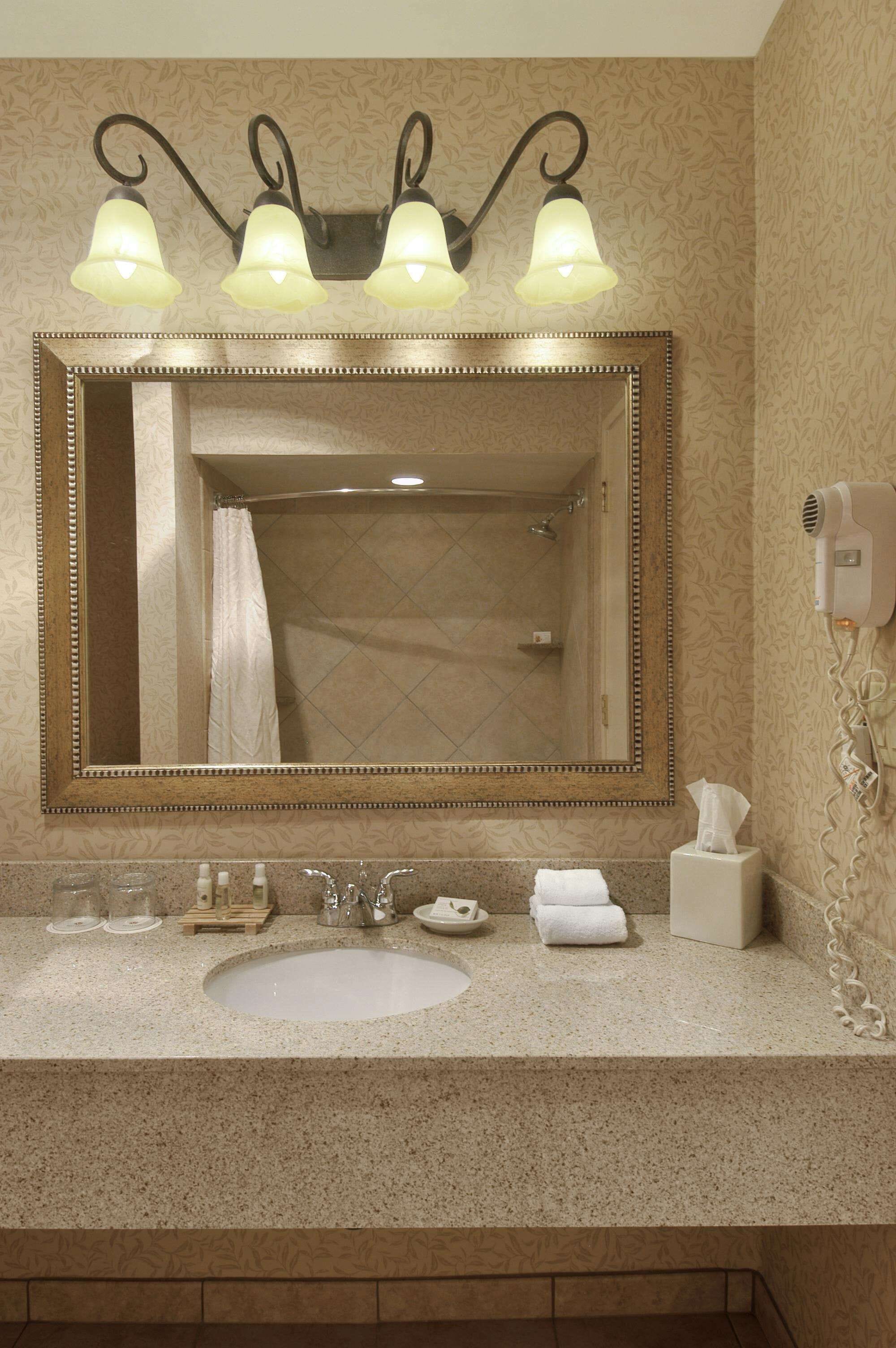 Une salle de bain avec un grand miroir, trois appliques murales, un lavabo, un porte-serviettes et un sèche-cheveux fixé au mur.