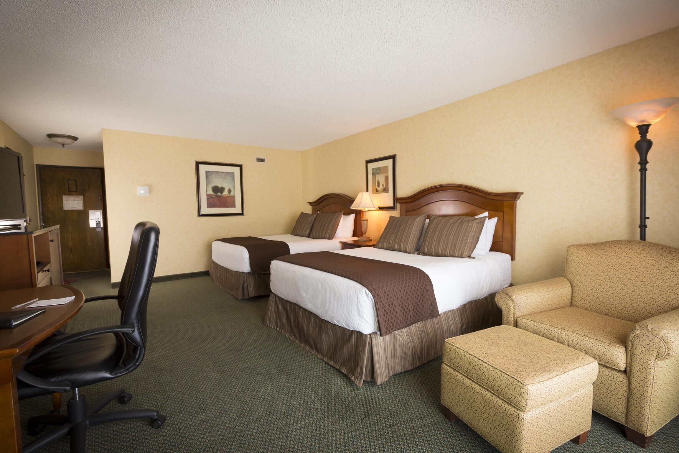 A hotel room with two beds, a desk, a chair, and a lamp.