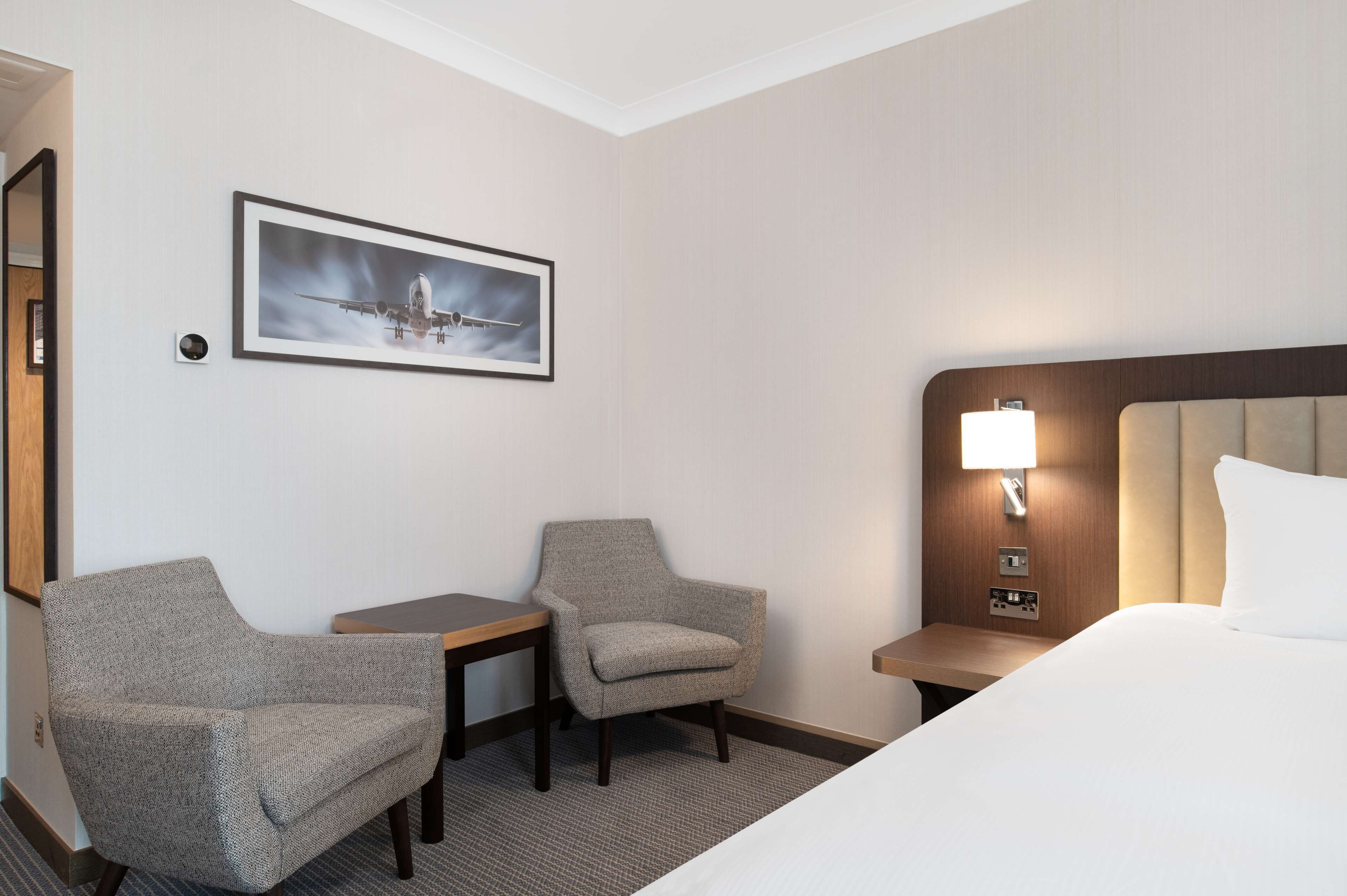 Ett hotellrum med en säng, två fåtöljer, ett litet bord, ett nattduksbord med en lampa och en inramad tavla på väggen.